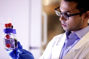 A man holds an electrochemical cell, which looks like two small bottles attached to each other, used for catalyst testing.